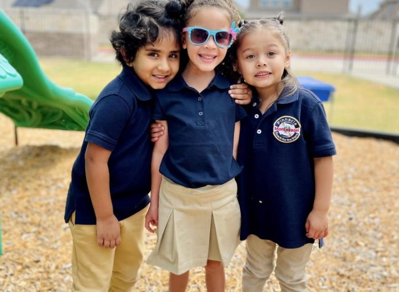 Three cheerful children smiling and posing together outdoors on the playground at Pinnacle Montessori of Castle Hills.